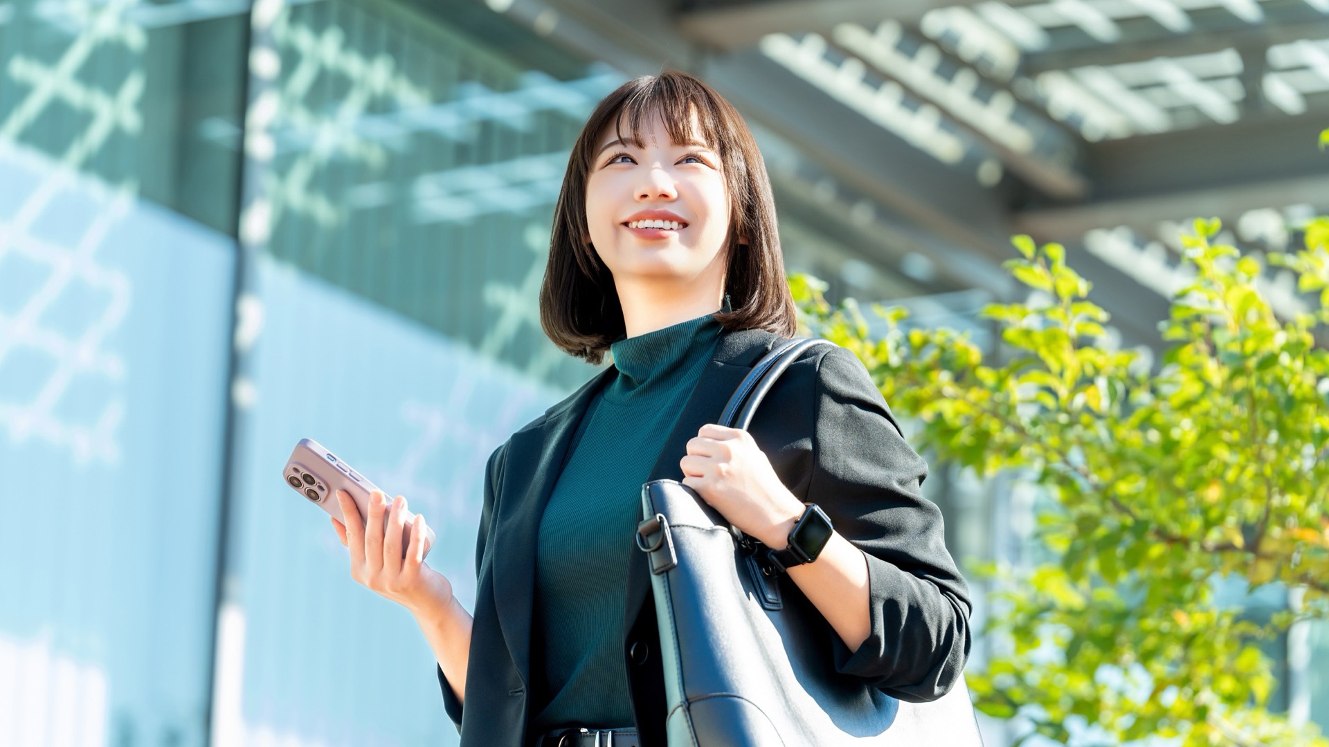 スマートフォンを手に、明るいオフィス街で笑顔を見せる女性